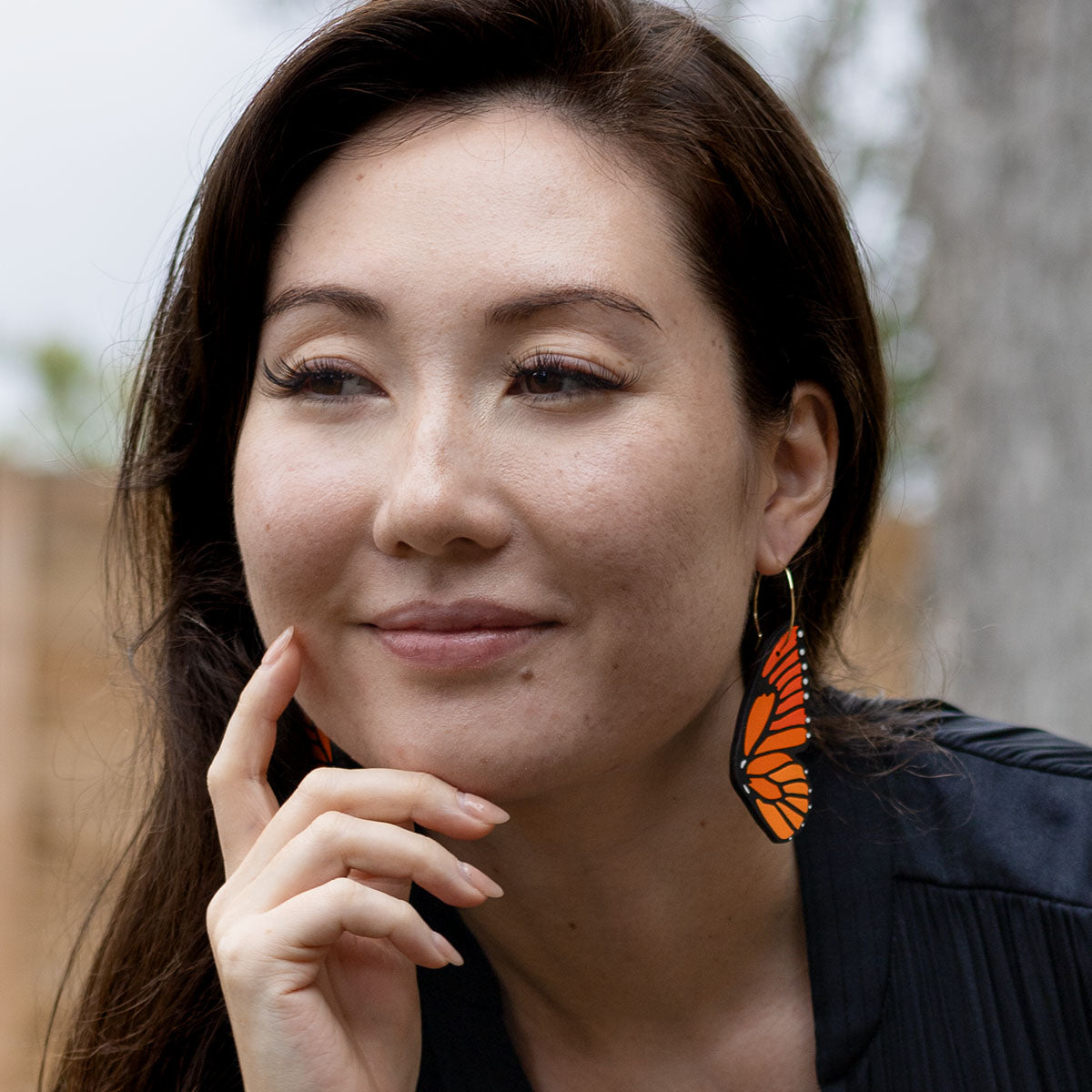 Woman wearing hand-painted monarch butterfly wing hoop earrings, resting her chin on her hand with a soft smile.