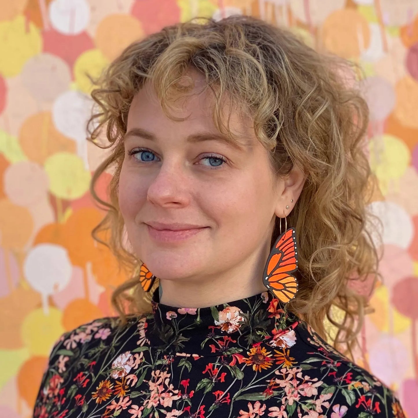 Woman wearing hand-painted monarch butterfly hoop earrings, standing in front of a colorful abstract mural, wearing a floral top.