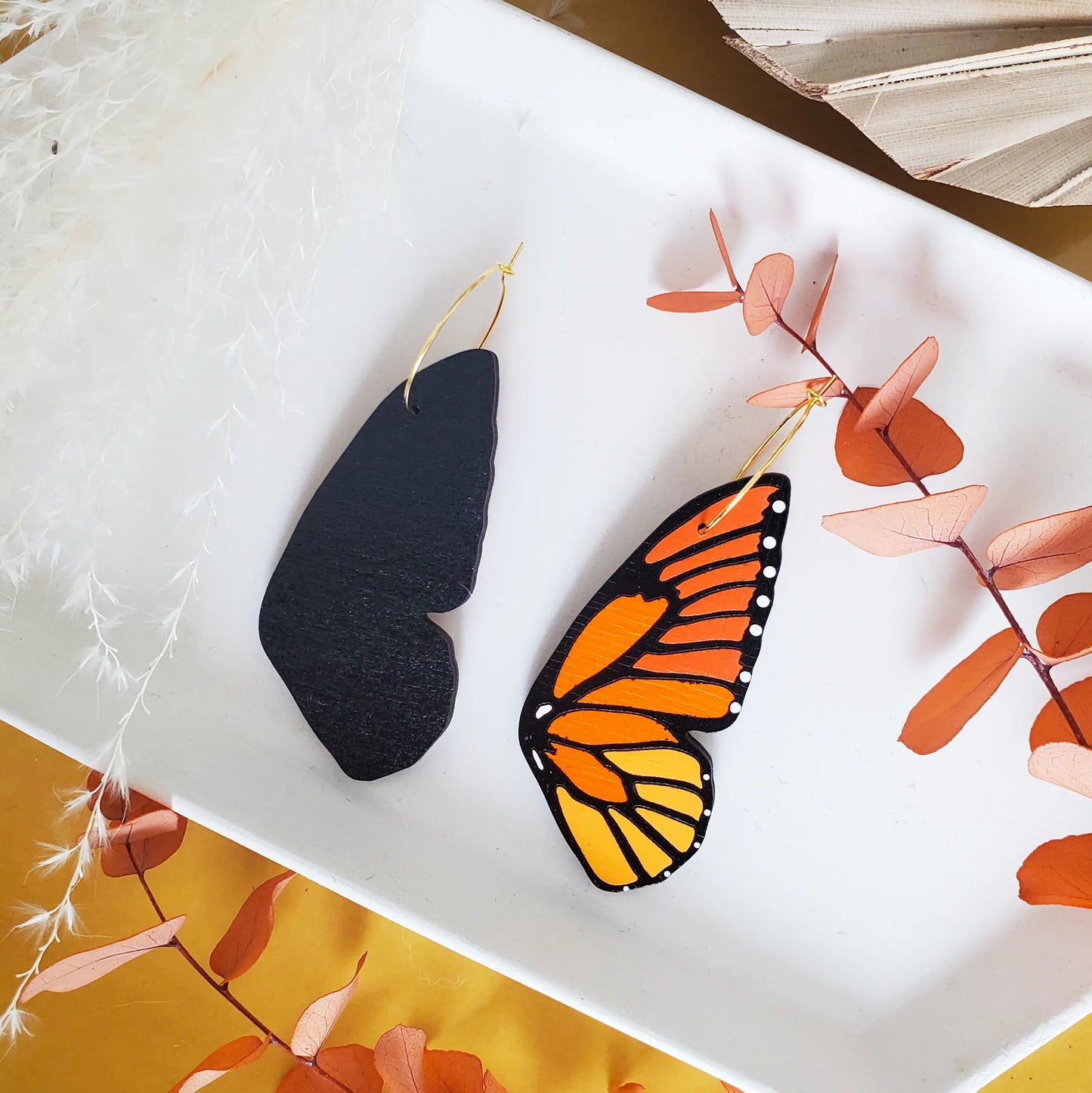 Front and back view of hand-painted monarch butterfly wing hoop earrings displayed on a white tray with orange and neutral botanical accents.