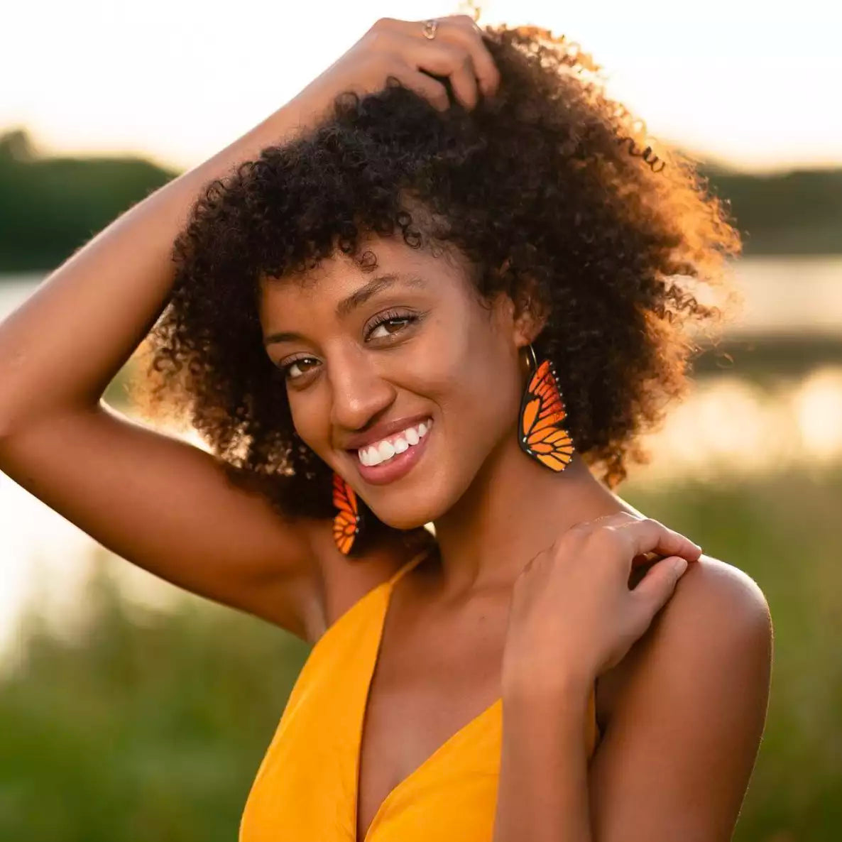Woman wearing hand-painted monarch butterfly wing hoop earrings in vibrant orange and black, smiling outdoors at sunset.