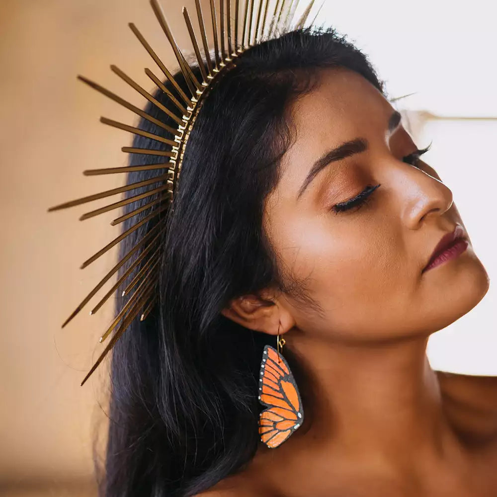 Woman wearing hand-painted monarch butterfly wing hoop earring and a gold sunburst crown, with eyes closed in a serene pose.