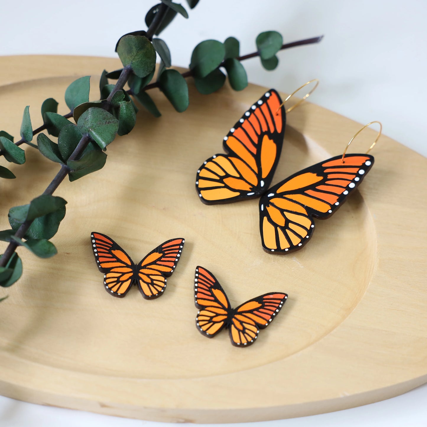 Set of hand-painted monarch butterfly earrings on a wooden tray, including large wing hoop earrings and smaller butterfly stud earrings with a eucalyptus branch.