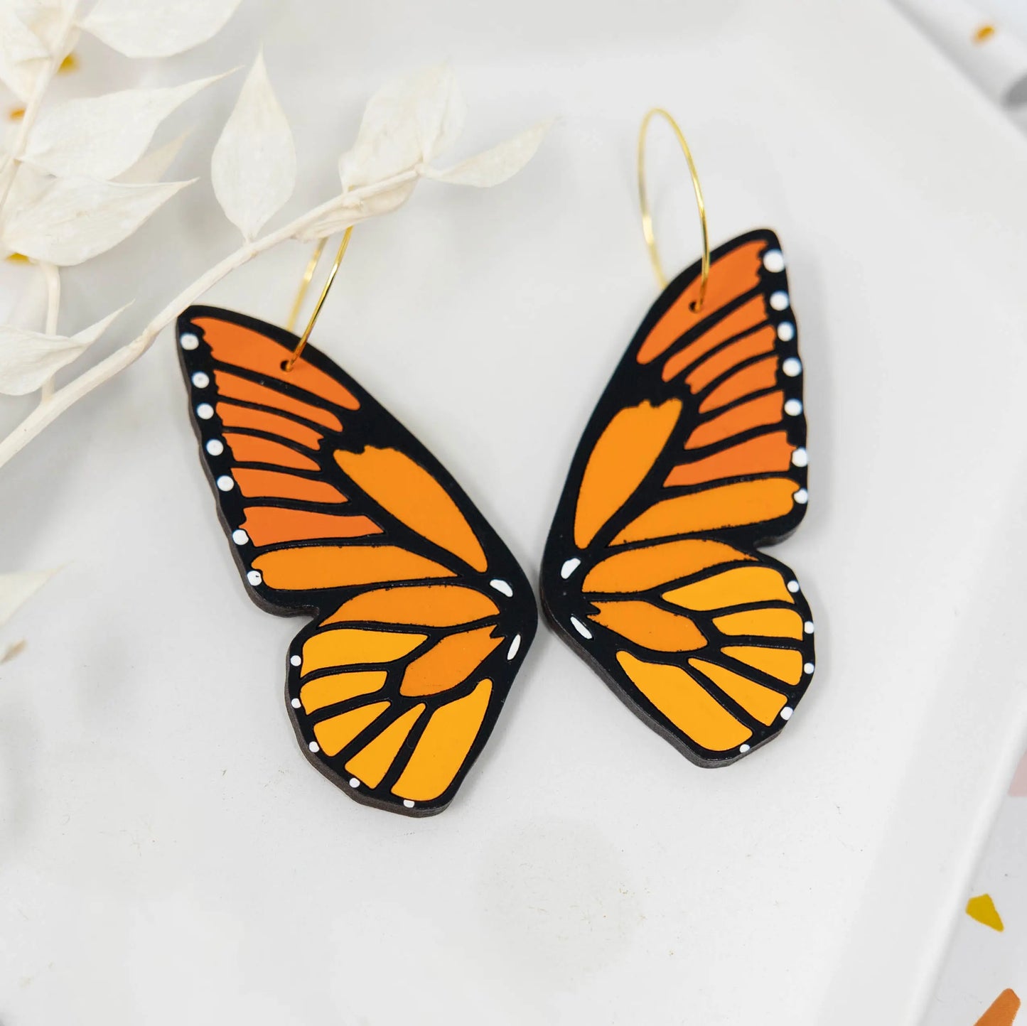 Pair of hand-painted monarch butterfly wing hoop earrings displayed on a white ceramic plate with dried white foliage.
