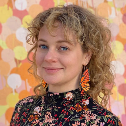 Woman wearing hand-painted monarch butterfly hoop earrings, standing in front of a colorful abstract mural, wearing a floral top.