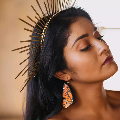 Woman wearing hand-painted monarch butterfly wing hoop earring and a gold sunburst crown, with eyes closed in a serene pose.